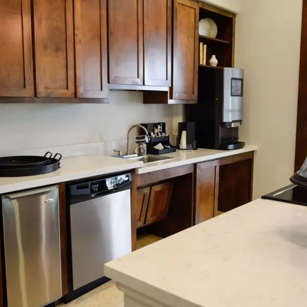 A modern kitchen with wooden cabinets, stainless steel appliances, and a white countertop. There's a large island in the foreground with a glass cloche, and a coffee maker and accessories can be seen on the counter.