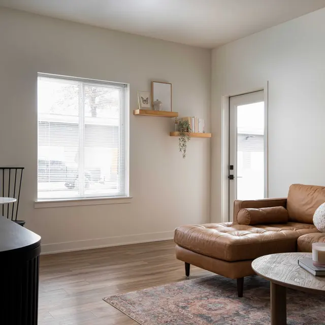 A cozy living room featuring a tan leather sectional sofa, a round coffee table, and a small dining area. There is a window with natural light and decorative shelves on the wall.