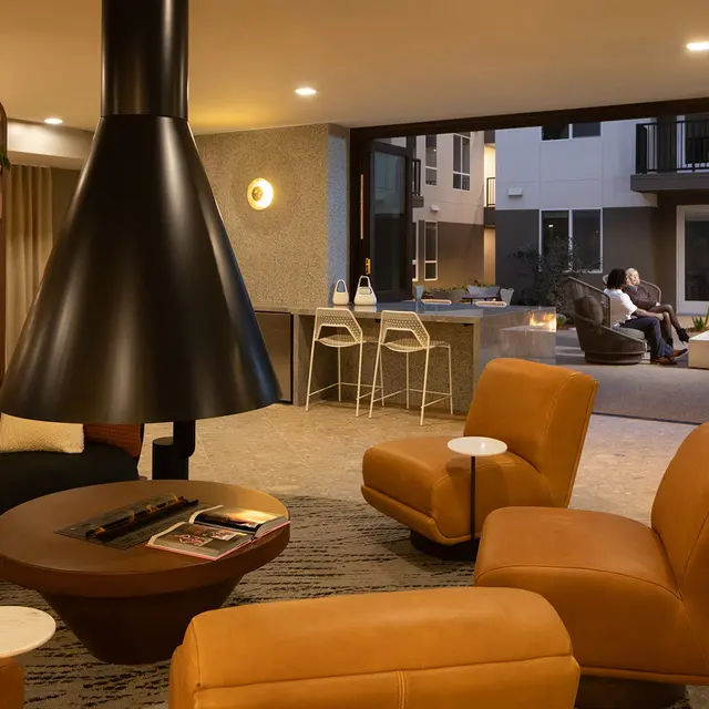 A modern amenity space with a black and orange color scheme, featuring a circular coffee table, two orange armchairs, and a sleek black fireplace. The background shows a patio area with two people sitting, illuminated softly at night.