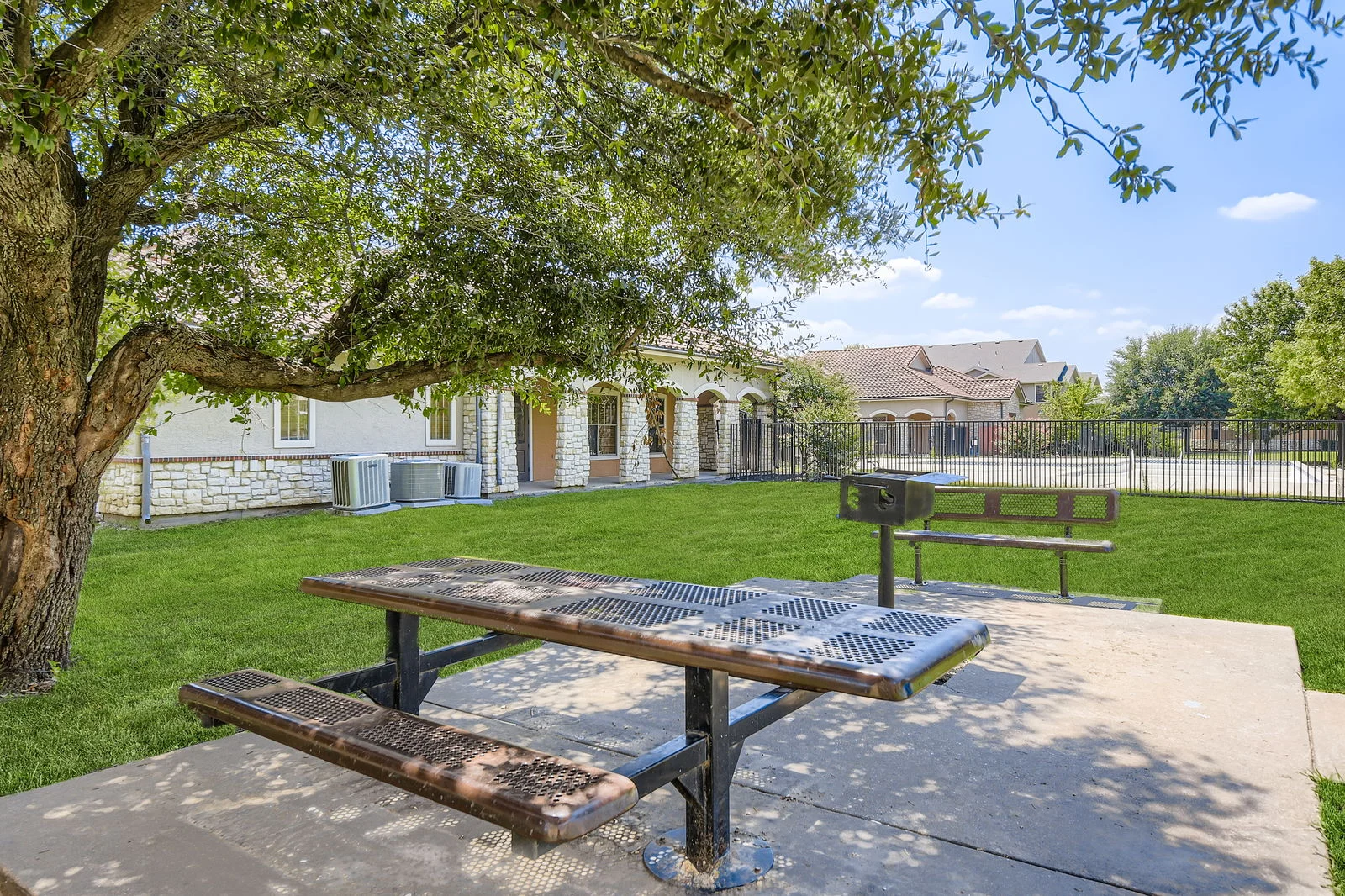 A picnic table under a tree in a grassy area with a grill nearby and a residential building in the background.