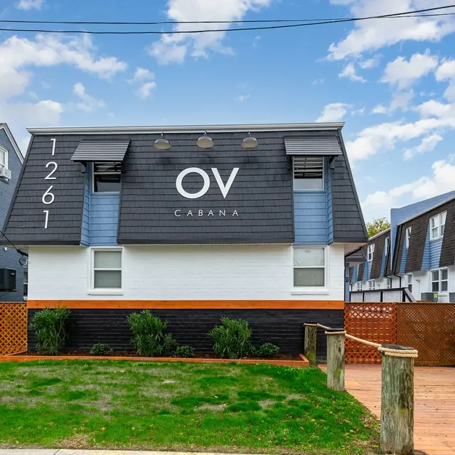 A multi-level building featuring a dark roof and a prominent sign saying 'OV CABANA'. The exterior is primarily white with some green grass and landscaping in the front.