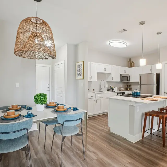 A modern kitchen and dining area featuring a dining table set for four with blue dishes and brown plates. The kitchen has white cabinetry, stainless steel appliances, and a large island with bar stools. The room is bright with a large pendant light overhead and natural light coming from the windows.