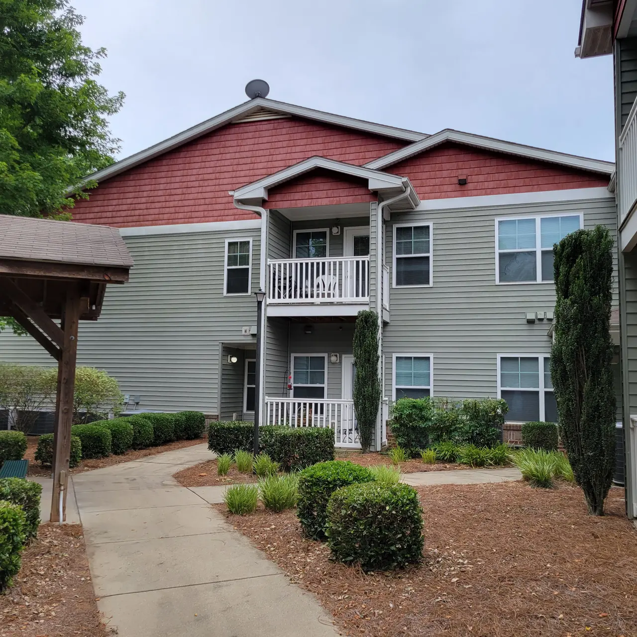 View of a multi-story apartment building with red and gray siding, surrounded by neatly landscaped greenery and pathways.