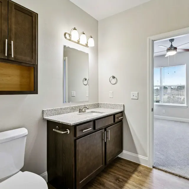 A modern bathroom featuring a toilet, a granite countertop with a sink, a large mirror, and overhead lighting. A doorway leads to a well-lit room with a window in the background.