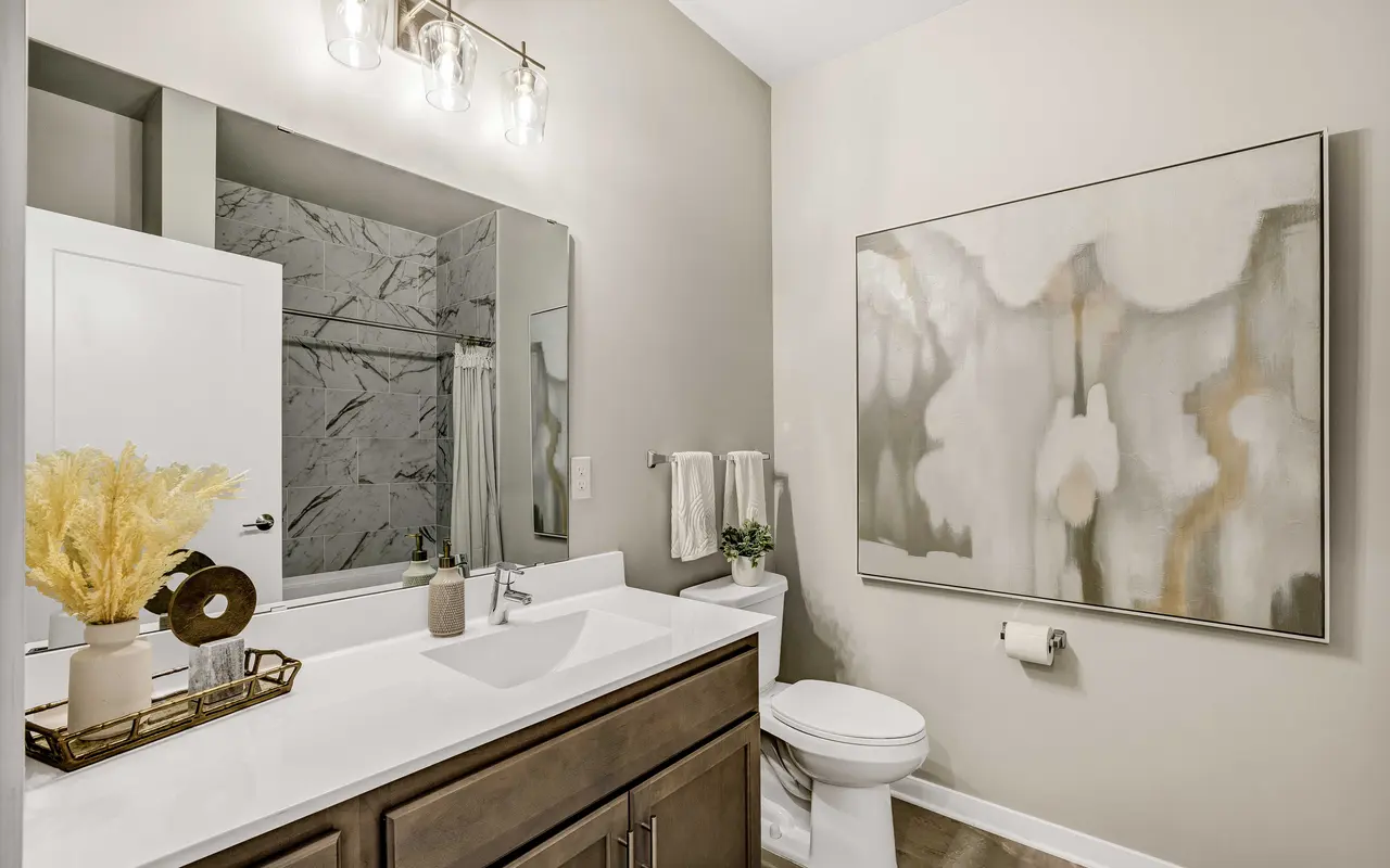 A modern bathroom featuring a large mirror, elegant lighting, a white countertop, and a minimalist design with a decorative painting and a plant.