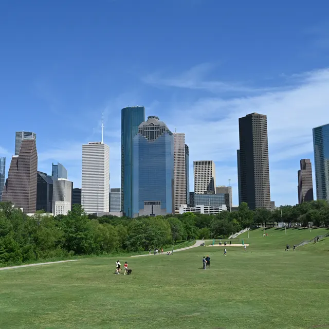 Market Square Park A panoramic view of the Houston skyline featuring tall skyscrapers against a clear blue sky, with a green park area in the foreground where people can be seen walking and enjoying the outdoors.