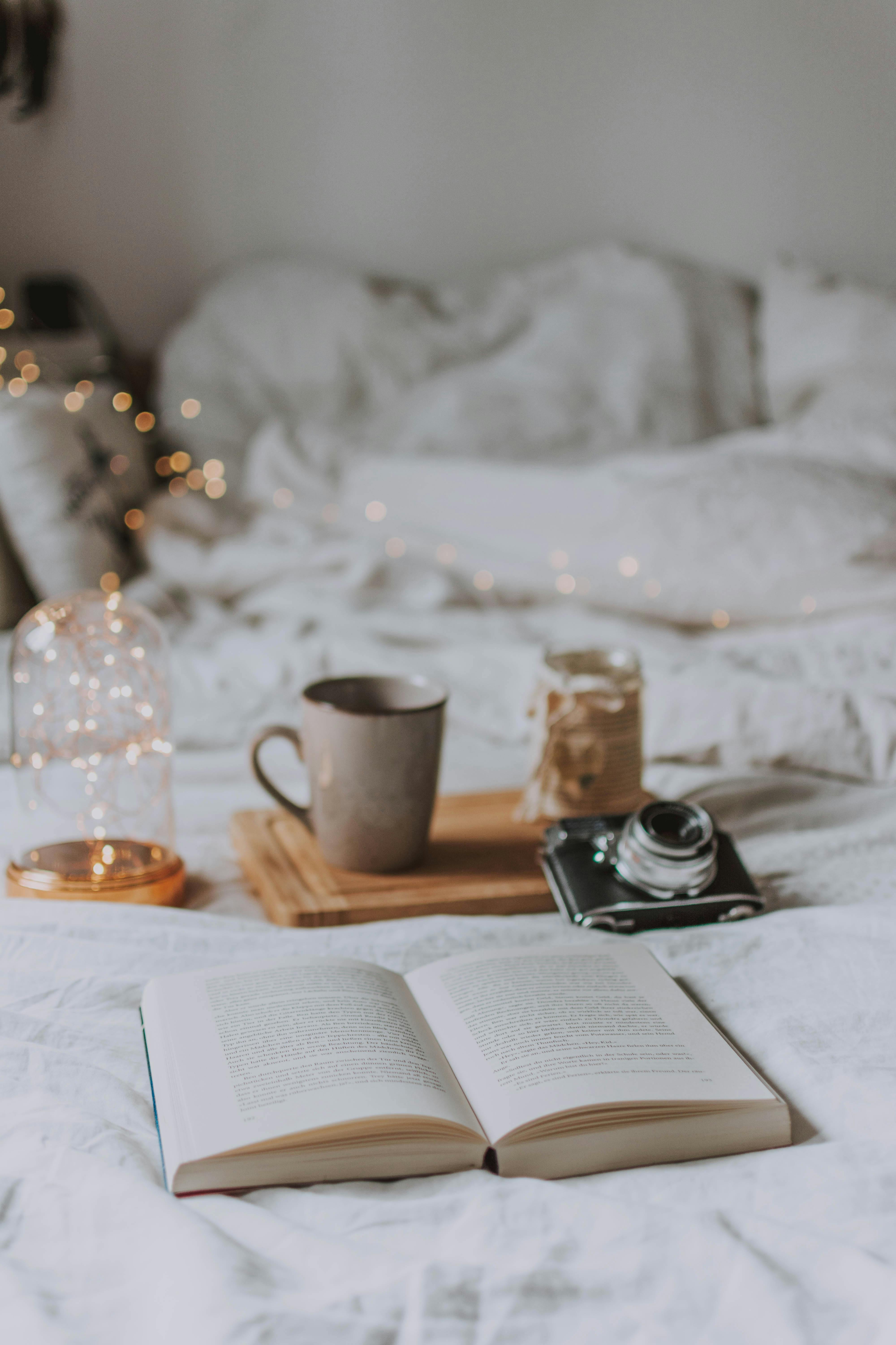 An open book lays on a bed with soft linens, accented by warm fairy lights, a mug of hot beverage, a jar, and a vintage camera on a wooden tray.