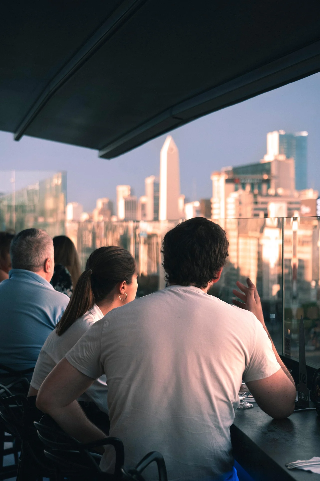 A group of people sitting at a bar, enjoying a panoramic view of a city skyline during sunset.