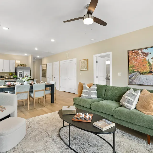 A modern living room featuring a green sofa, coffee table with decorative items, and a dining area with a wooden table set for meals. The room has large windows and a painting depicting a path lined with trees in autumn colors.