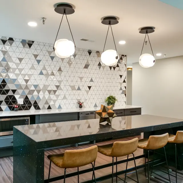 A modern kitchen featuring a geometric tiled backsplash in black, white, and gray tones. The kitchen island is topped with a dark countertop and has brown bar stools arranged around it. Pendant lights hang above the island, illuminating the space.