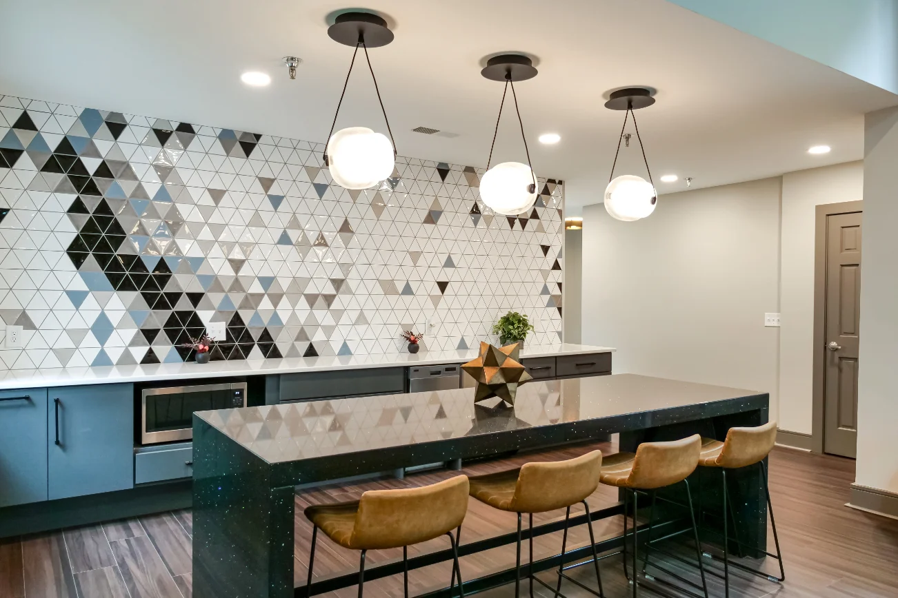 A modern kitchen featuring a geometric tiled backsplash in black, white, and gray tones. The kitchen island is topped with a dark countertop and has brown bar stools arranged around it. Pendant lights hang above the island, illuminating the space.
