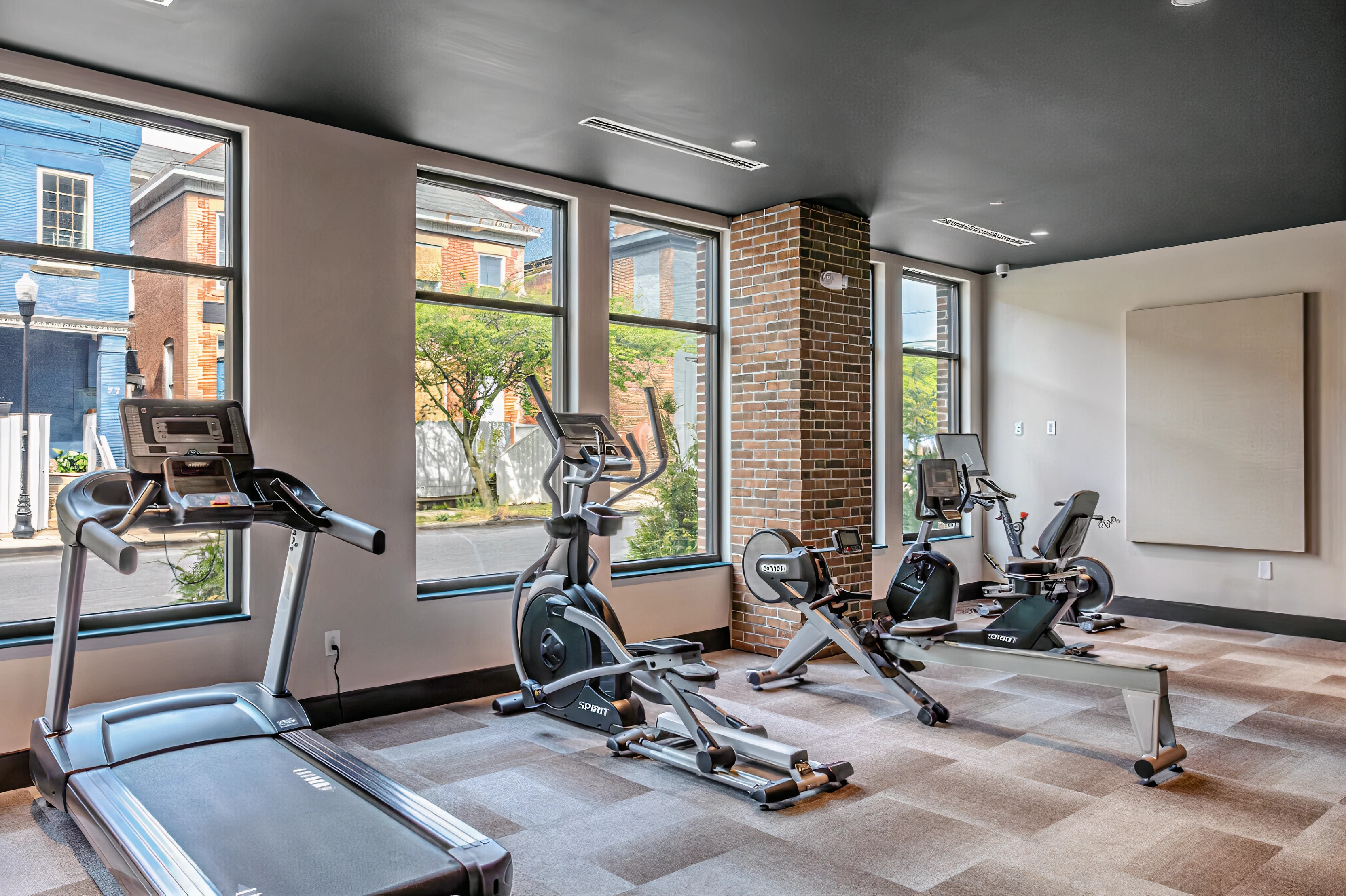 An interior view of a modern gym with various exercise equipment, including a treadmill, elliptical machine, and stationary bike, situated by large windows that provide natural light.