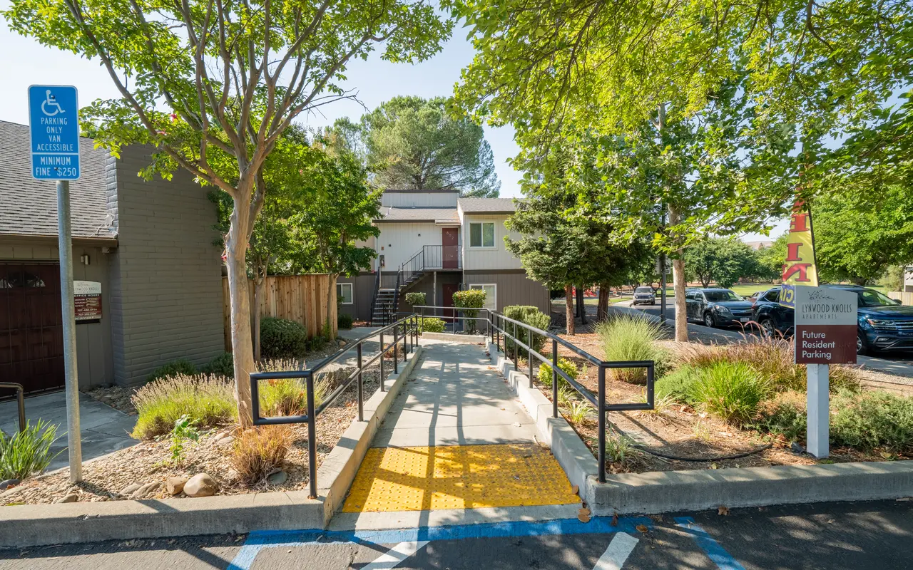 An accessible pathway leading to a building, with ramped entry and markings for visually impaired pedestrians, surrounded by trees and foliage.