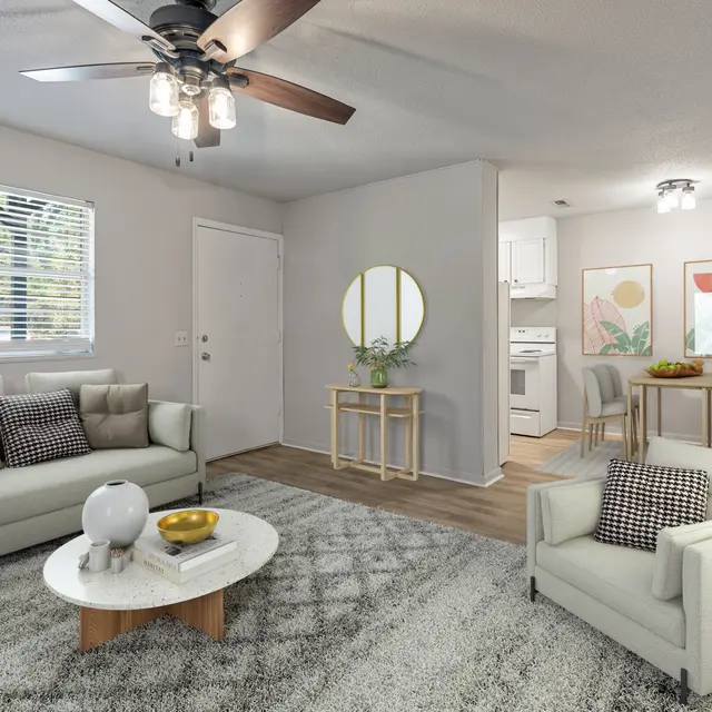 A spacious and modern living room featuring a comfortable gray sofa with decorative cushions, a stylish cream-colored armchair, a round wooden coffee table, and a light gray rug. There's a round mirror on the wall with a small plant beside it. A bright kitchen area is visible in the background, alongside a dining nook with light-colored chairs and artwork on the walls.