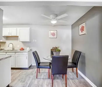 A modern dining area featuring a round glass table with four black chairs. The room is well-lit with a ceiling fan and has a neutral color scheme, including a dark accent wall with framed floral artwork. The kitchen is visible in the background, showcasing white cabinets and a clean countertop.