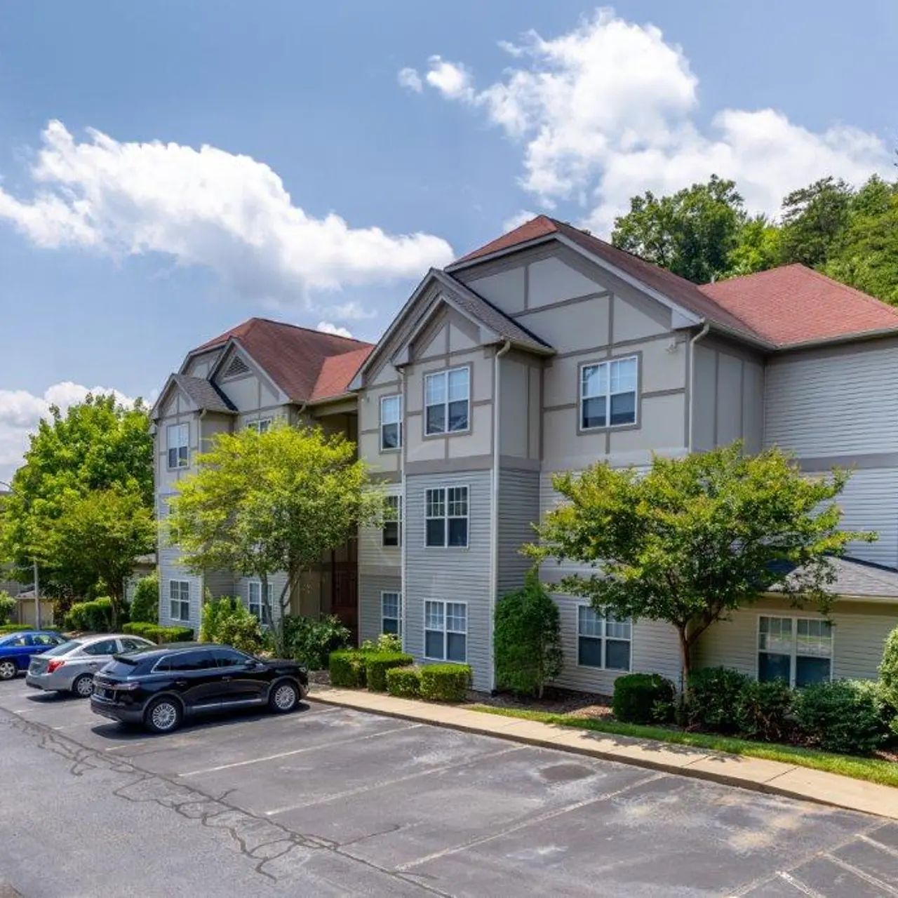 Exterior view of a multi-story apartment complex surrounded by trees and parked cars on a sunny day.