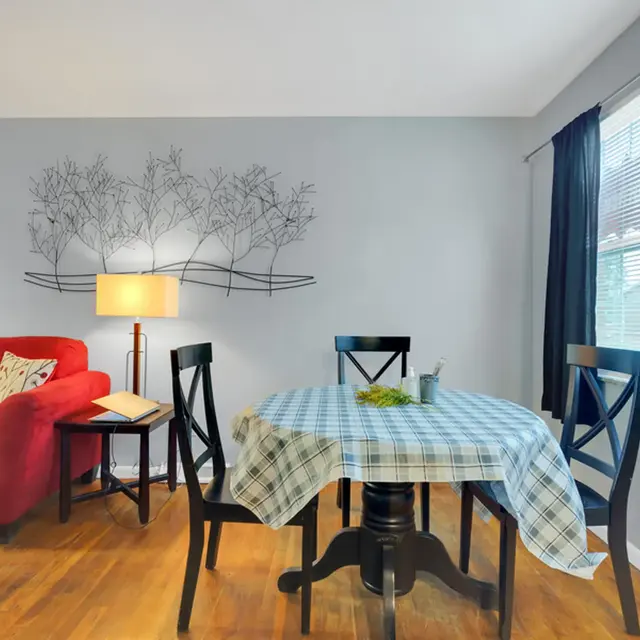 A cozy dining area featuring a round table dressed in a blue checkered tablecloth, surrounded by four black chairs. To the left, a red sofa with a decorative pillow complements the space. A floor lamp with a light shade stands next to the table, and a wall decoration of trees adds a serene touch. Natural light filters in through a window dressed with dark curtains.