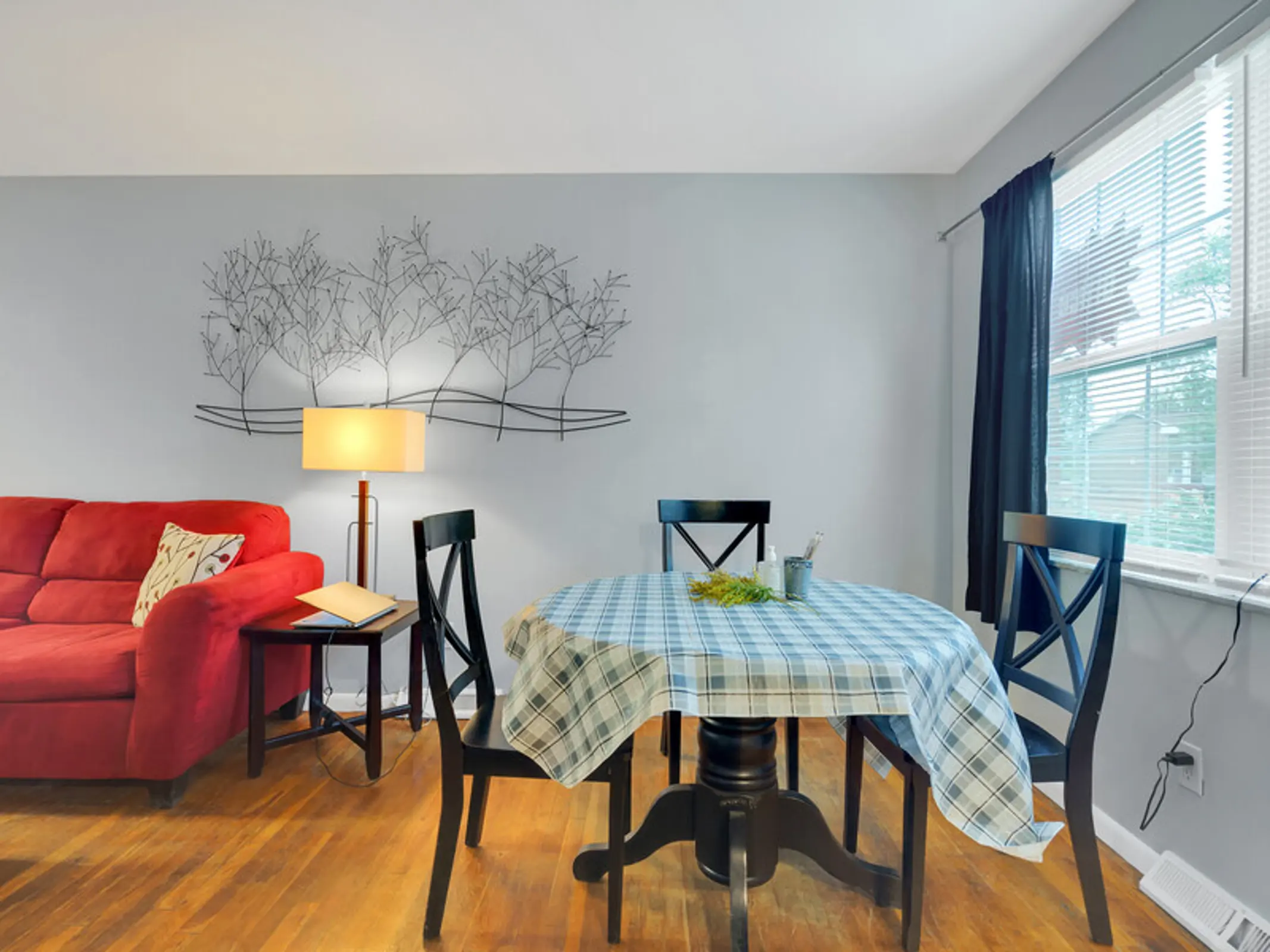 Modern Dining Area A cozy dining area featuring a round table dressed in a blue checkered tablecloth, surrounded by four black chairs. To the left, a red sofa with a decorative pillow complements the space. A floor lamp with a light shade stands next to the table, and a wall decoration of trees adds a serene touch. Natural light filters in through a window dressed with dark curtains.