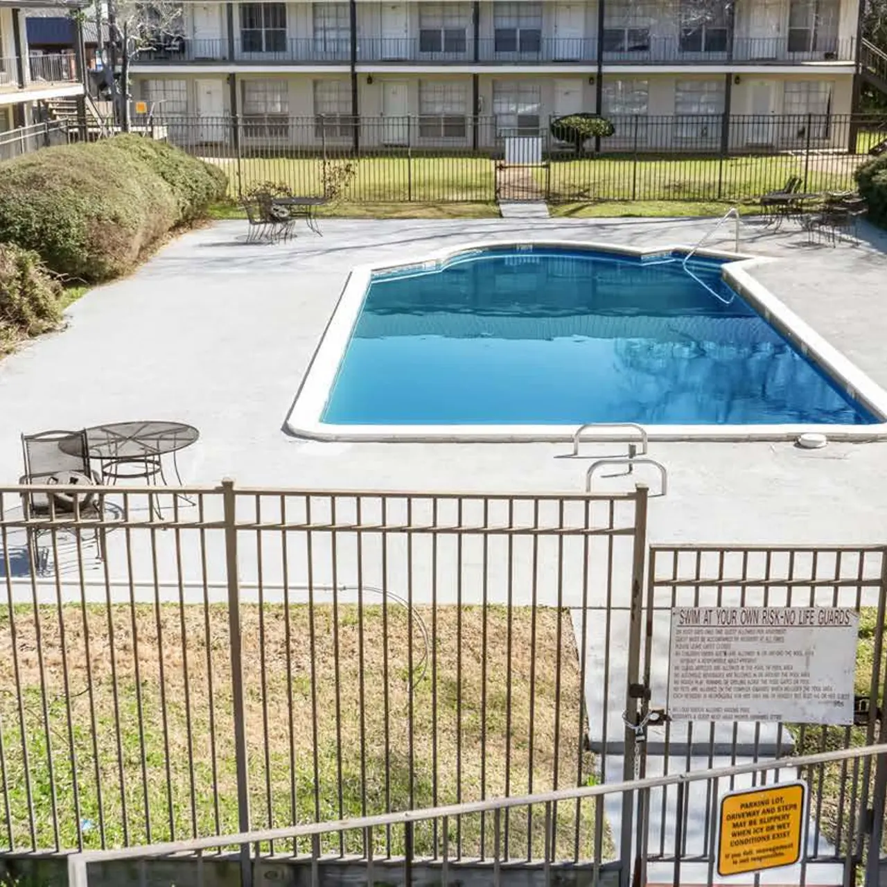 A fenced swimming pool area surrounded by greenery and several apartment buildings in the background.