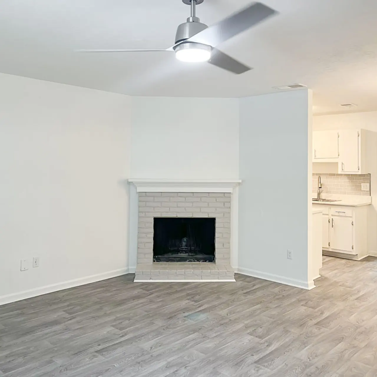 A spacious living room featuring light gray flooring and a white brick fireplace. The walls are mostly white, enhancing the brightness of the room. A ceiling fan is mounted overhead, and a doorway leads to a kitchen area that can be seen in the background.