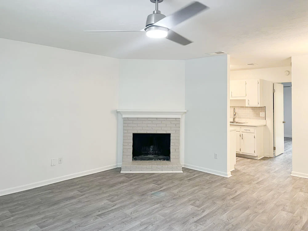 A spacious living room featuring light gray flooring and a white brick fireplace. The walls are mostly white, enhancing the brightness of the room. A ceiling fan is mounted overhead, and a doorway leads to a kitchen area that can be seen in the background.