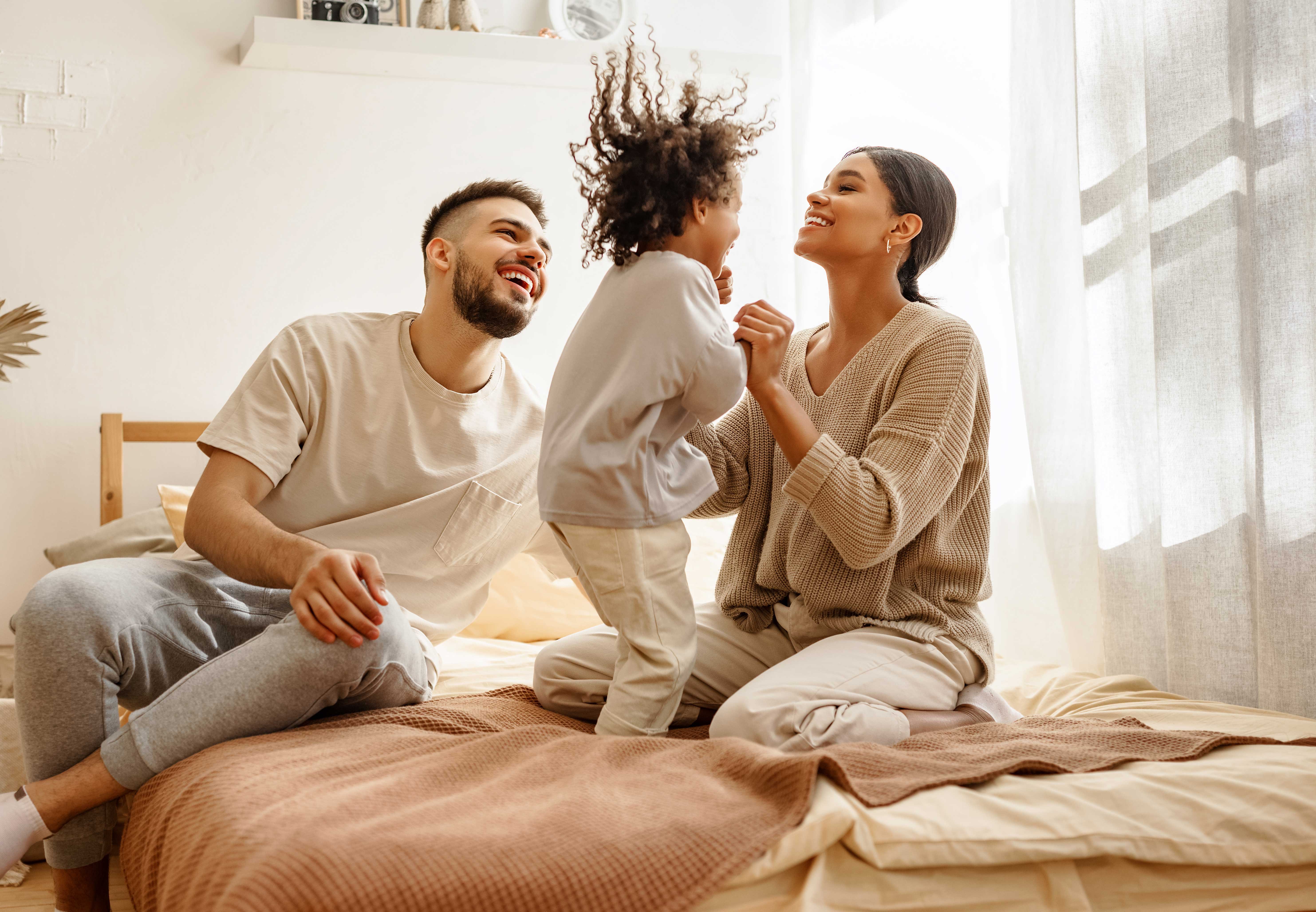 A happy family playing together in a cozy bedroom. A child with curly hair is being playfully engaged by two adults, a man and a woman, on a bed with soft bedding.