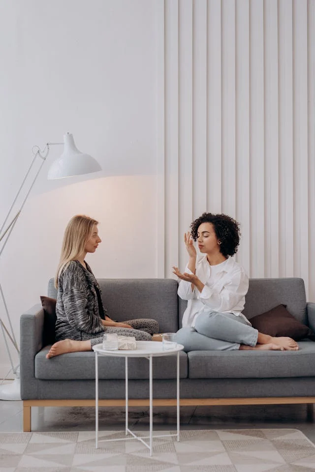 Women Engaging in Conversation Two women sitting on a sofa engaged in conversation, with one gesturing and the other listening. A small table with items is placed in front of them.