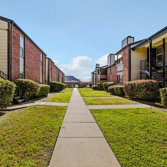 A pathway leading through an apartment complex, lined with greenery and facing buildings on either side.