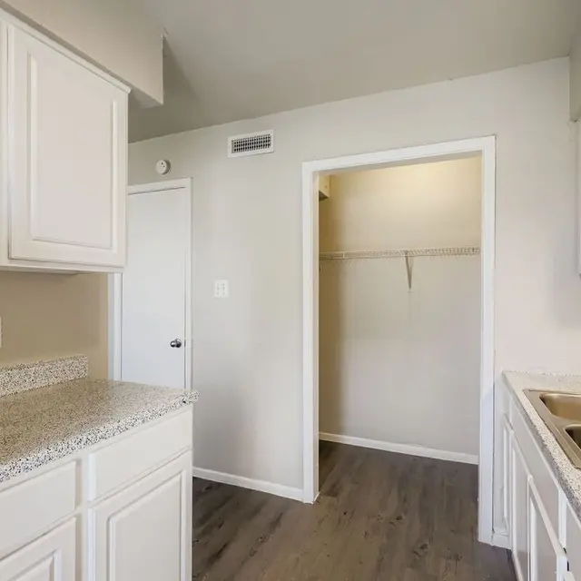 A modern kitchen with white cabinets and a granite countertop, featuring a stainless steel sink and an entryway leading to a closet area.