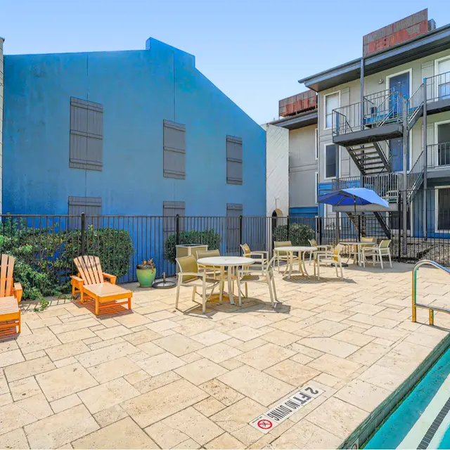 Apartment Pool Area A sunny pool area with lounge chairs and tables, surrounded by fencing and a blue building in the background.