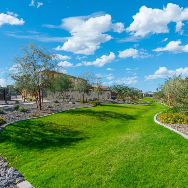 A picturesque view of a landscaped park that features a curved grassy area bordered by rocks and shrubs under a bright blue sky with fluffy white clouds.