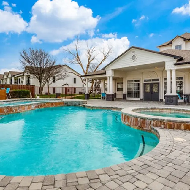 A beautiful outdoor pool area with a hot tub, surrounded by cobblestone, and backed by residential buildings. Bright blue sky with fluffy clouds add to the pleasant ambiance.
