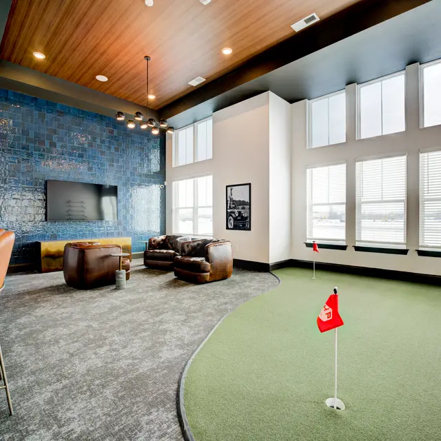A modern lounge space featuring a putting green, comfortable seating, and large windows allowing natural light to enter.