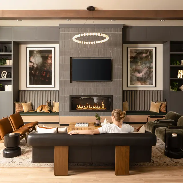 A modern living room featuring a cozy arrangement with a large couch, armchairs, and a fireplace. The room has large windows allowing natural light, and decorative shelves with plants and artwork.