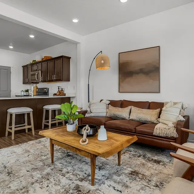 A cozy modern living room featuring a brown leather sofa, wooden coffee table, and a small kitchenette in the background with bar stools. Brightly lit with recessed lighting and a large piece of artwork on the wall.