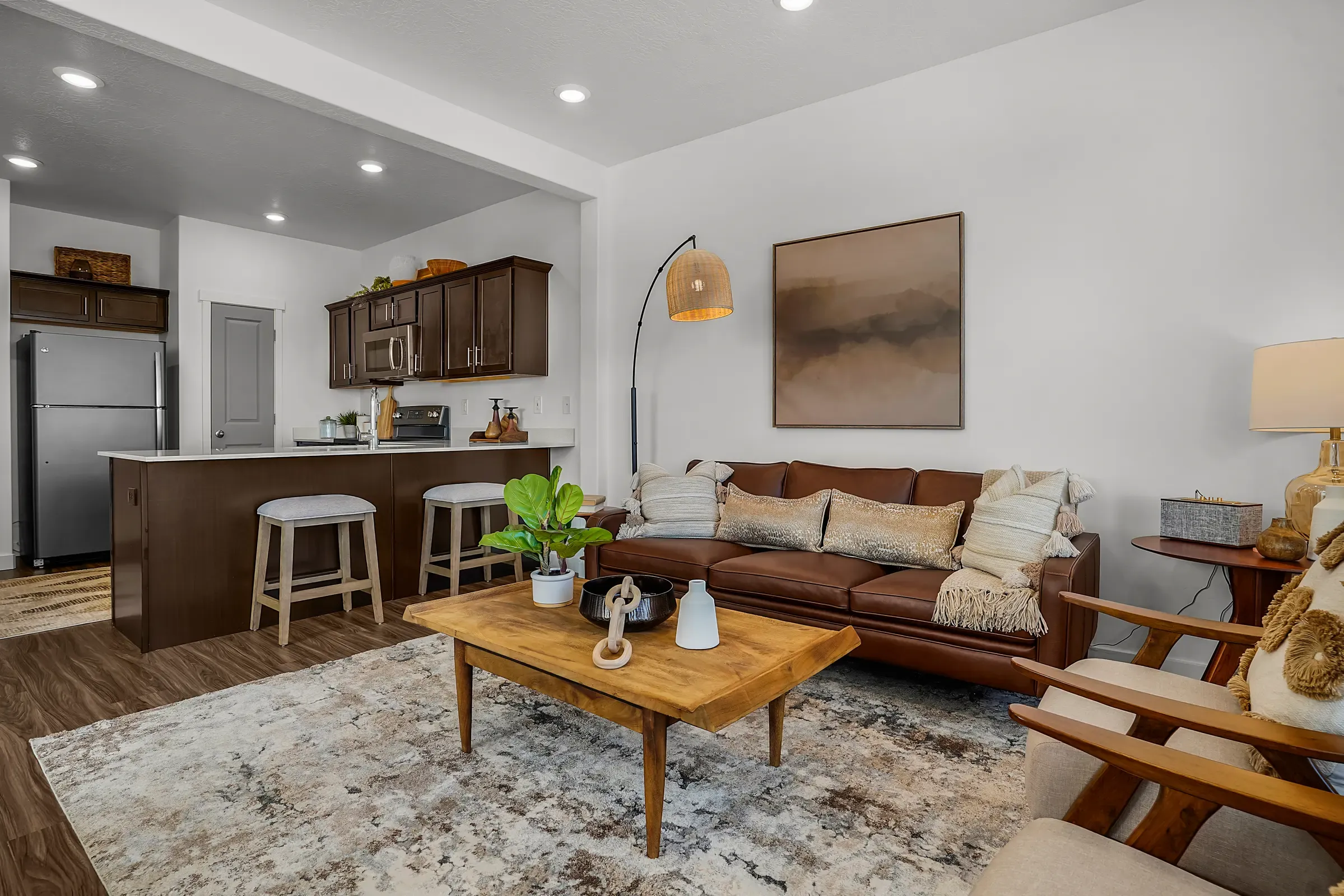 A cozy modern living room featuring a brown leather sofa, wooden coffee table, and a small kitchenette in the background with bar stools. Brightly lit with recessed lighting and a large piece of artwork on the wall.