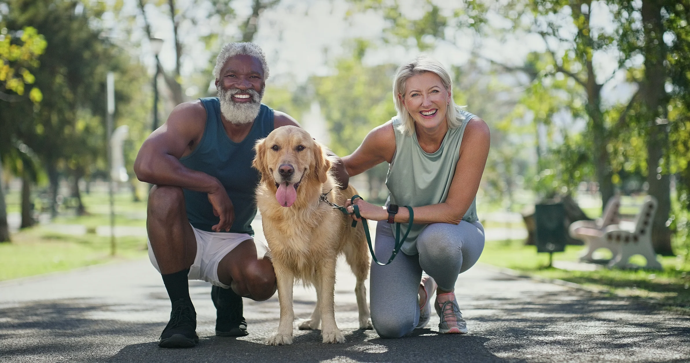 Happy Moments with a Dog in the Park A man and a woman kneeling beside a golden retriever in a park setting. The man has a beard and is wearing a tank top and shorts, while the woman is in a sport top and leggings. They both are smiling at the camera, surrounded by trees and sunlight.