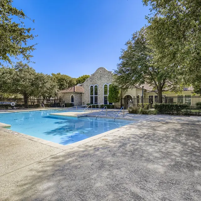 A clear blue swimming pool surrounded by a concrete deck and lush greenery, with a building in the background featuring decorative arches and large windows.