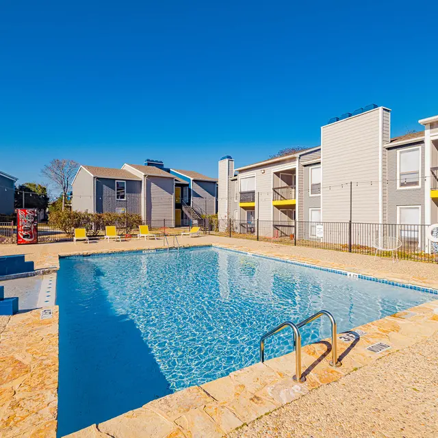View of a clear blue swimming pool surrounded by apartment buildings under a bright blue sky.