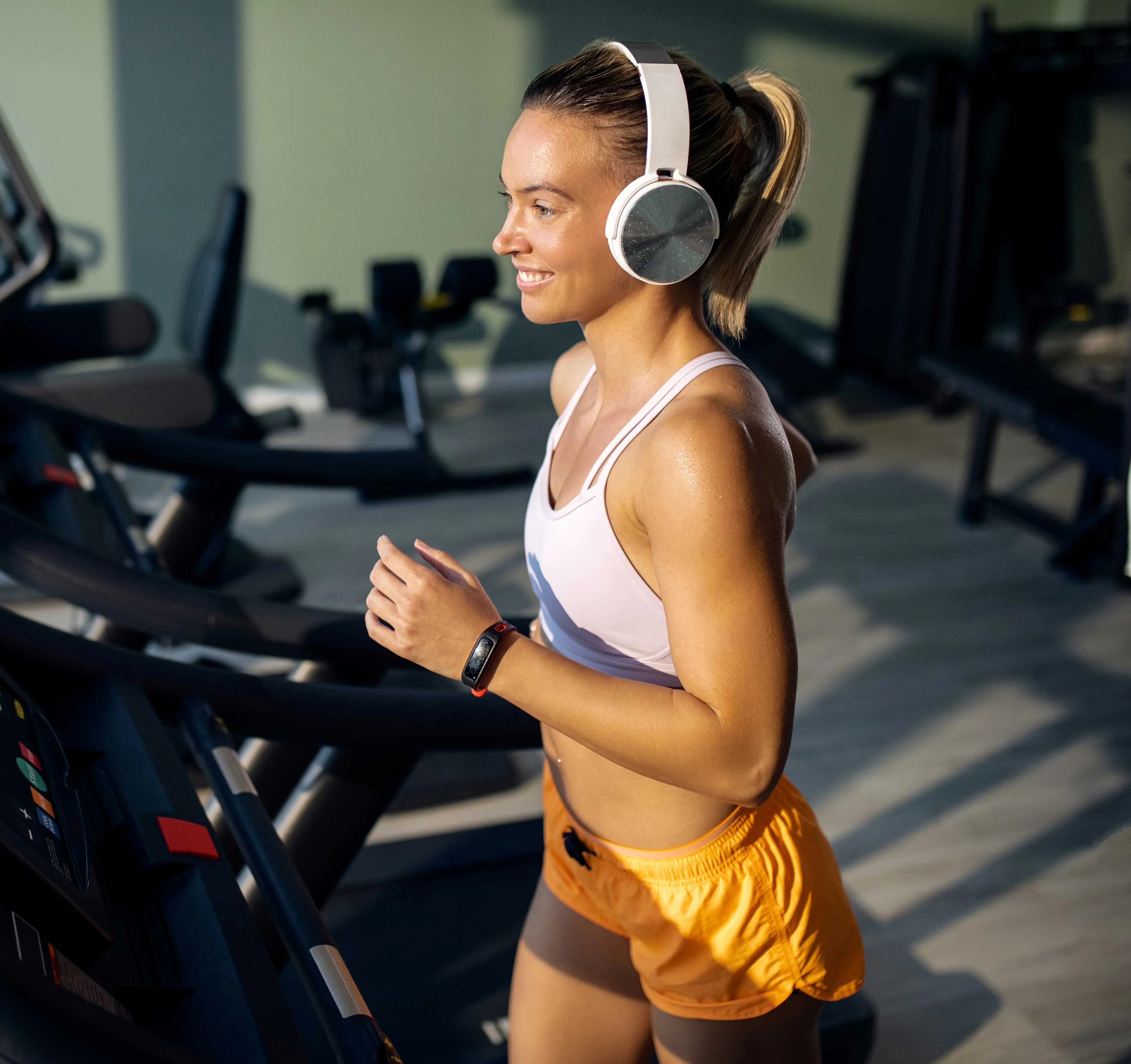 A young woman jogging on a treadmill, wearing headphones and workout attire, smiling as she exercises in a gym setting.