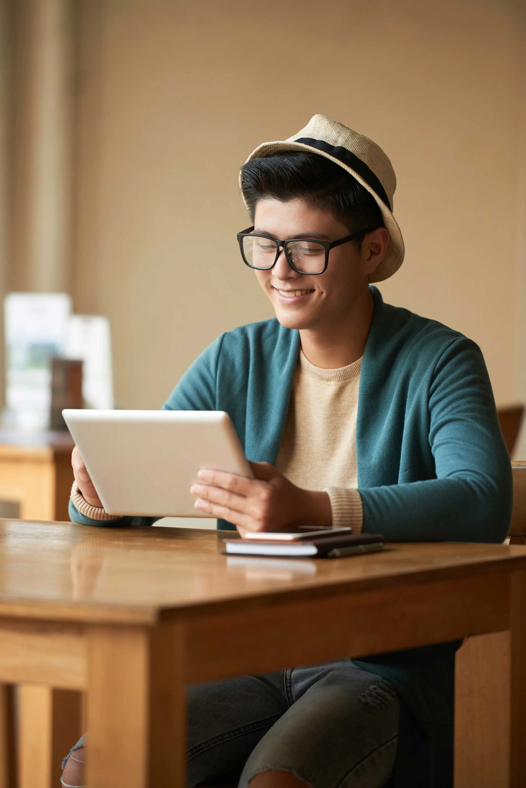 Young Man Using Tablet A young man with glasses and a hat sitting at a wooden table, looking at a tablet with a smile. He is wearing a blue cardigan and a beige shirt, with a notebook and phone on the table.