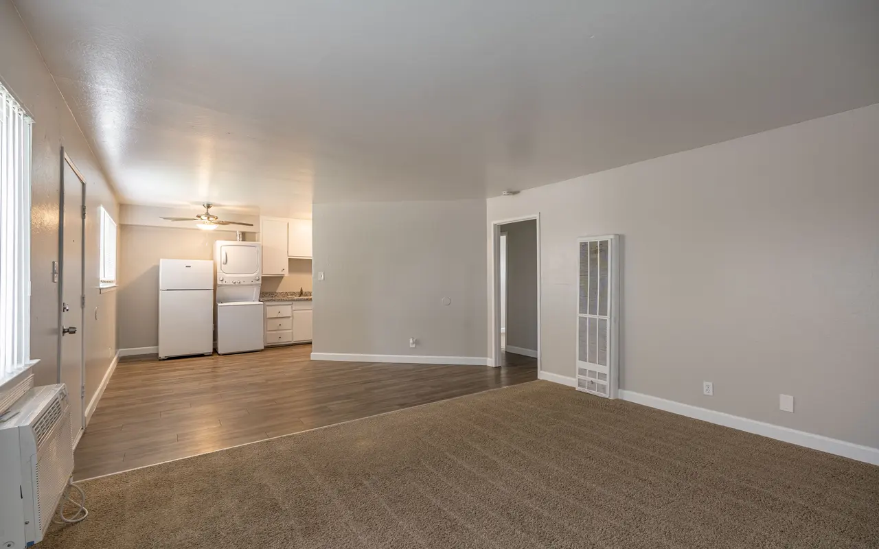 A spacious living area of an apartment with a kitchen visible in the background. The flooring is a mix of carpet and laminate, with white walls and bright lighting.
