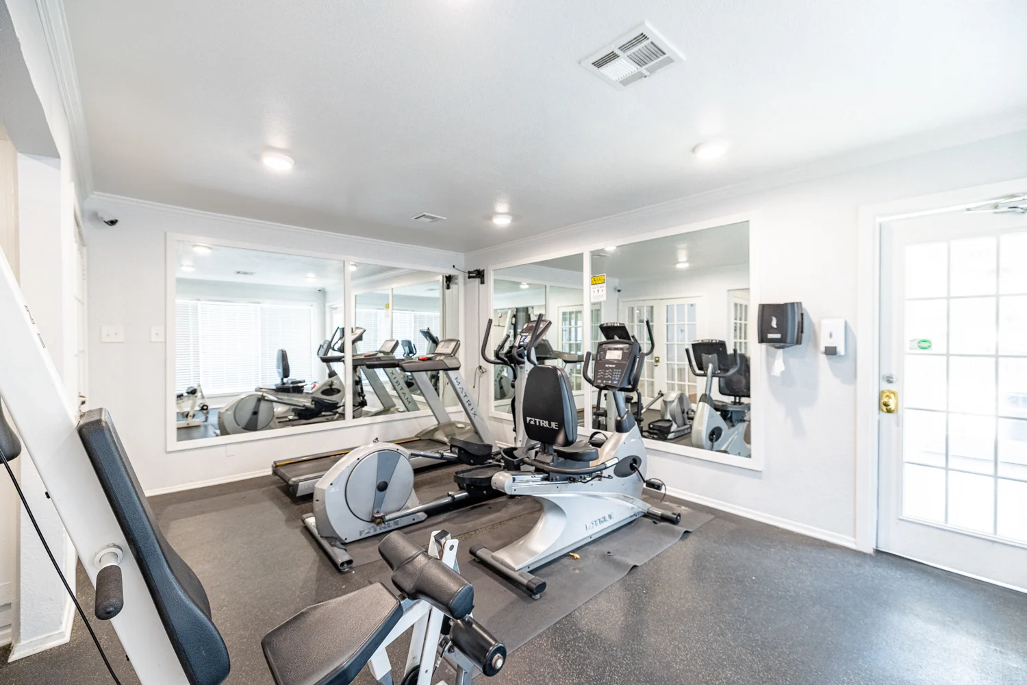 Modern Gym Interior An interior view of a gym with exercise equipment including treadmills, an elliptical machine, and a stationary bike. Mirrors line one wall and there's a door leading outside.