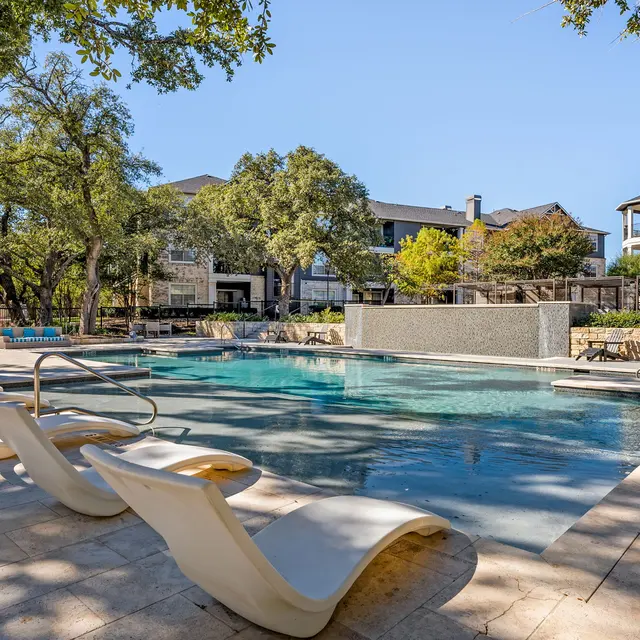 A tranquil swimming pool area surrounded by trees and lounge chairs under the sun.