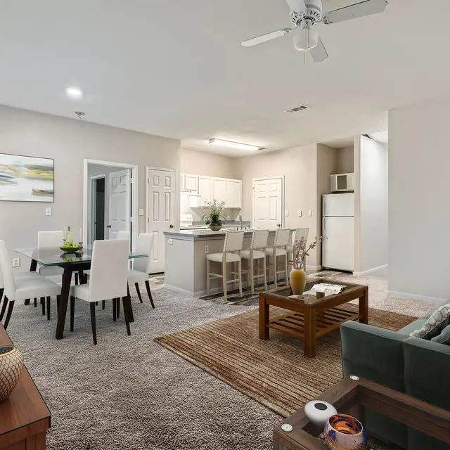A spacious modern apartment interior featuring a living area with a green couch, a wooden coffee table, and a television. The dining area has a glass table with seating for six. In the background, there's a kitchen with white cabinetry and appliances.