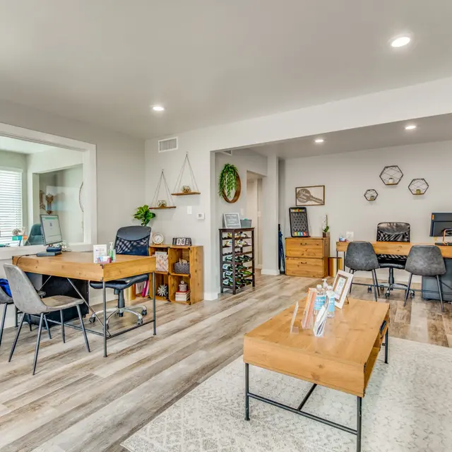 A modern office space with wooden flooring, featuring two desks with chairs on the left side and a seating area with a blue couch on the right side. The walls are light-colored, and there are decorative elements like plants and wall art.