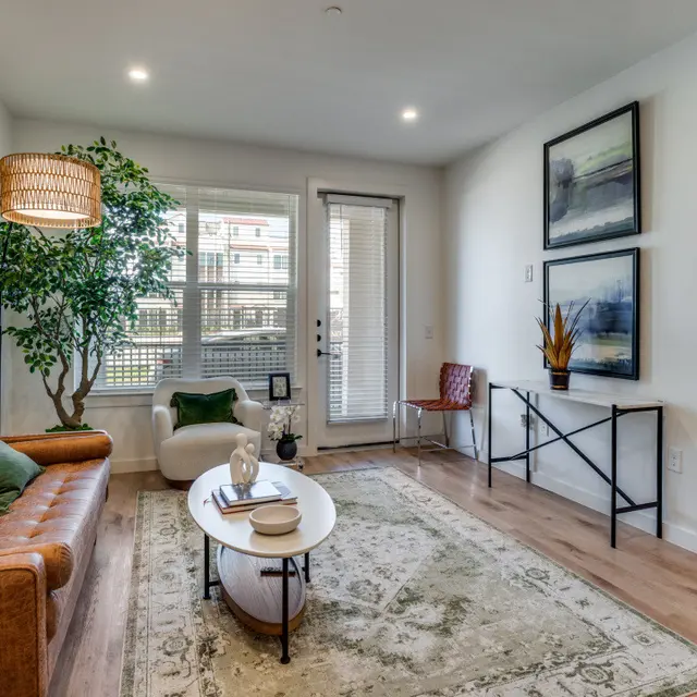 A modern living room featuring a brown leather sofa, a round coffee table, and a green plant, with large windows allowing natural light and artwork on the walls.