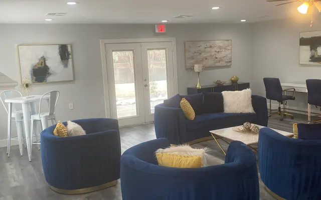 A cozy living area featuring plush navy blue circular chairs with gold accents, a light wooden table, and large windows allowing natural light. The walls are painted in a light gray color with abstract art pieces.