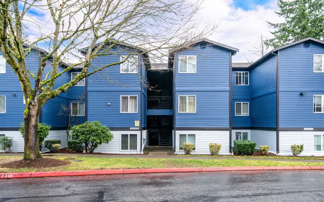 Exterior view of a blue apartment building with tree and shrubs in front.