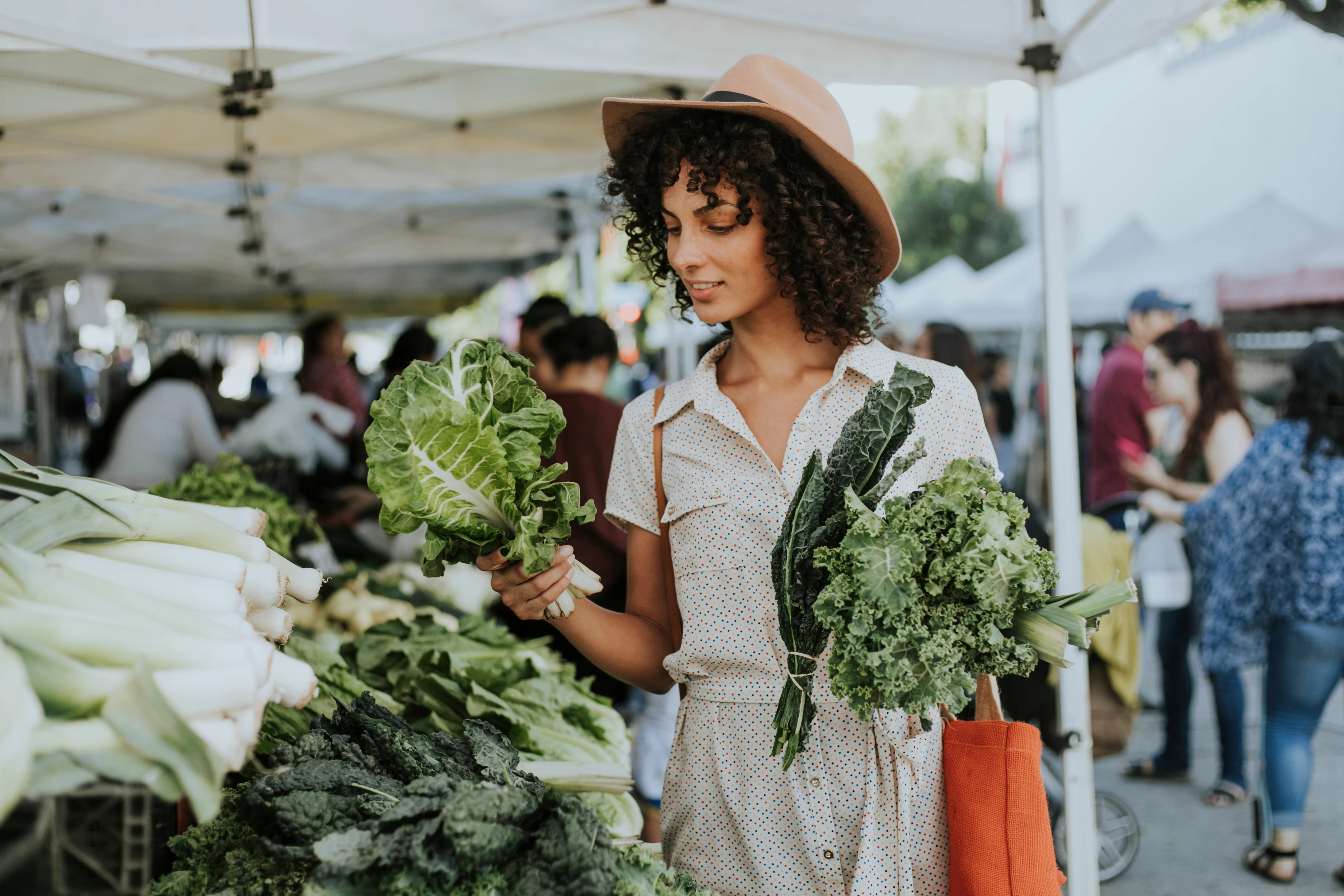Shopping for Fresh Produce at the Farmer's Market A woman in a summer dress and hat shopping for vegetables at a farmer's market, holding lettuce and kale while surrounded by other vendors and shoppers.